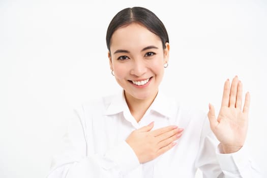 Friendly young businesswoman, asian lady raises one hand and puts palm on heart, makes promies, introduces herself, stands over white background.
