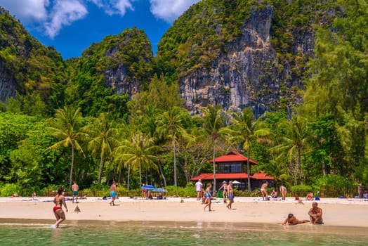 Bungalow house with red roof among coconut palms near the cliffs with people sunbathing and swimming in emerald water on Railay beach west, Ao Nang, Krabi, Thailand.