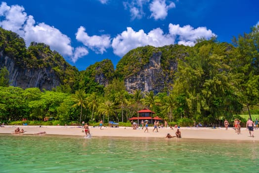 Bungalow house with red roof among coconut palms near the cliffs with people sunbathing and swimming in emerald water on Railay beach west, Ao Nang, Krabi, Thailand.