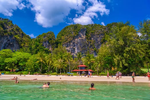 Bungalow house with red roof among coconut palms near the cliffs with people sunbathing and swimming in emerald water on Railay beach west, Ao Nang, Krabi, Thailand.