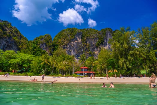 Bungalow house with red roof among coconut palms near the cliffs with people sunbathing and swimming in emerald water on Railay beach west, Ao Nang, Krabi, Thailand.