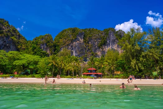 Bungalow house with red roof among coconut palms near the cliffs with people sunbathing and swimming in emerald water on Railay beach west, Ao Nang, Krabi, Thailand.