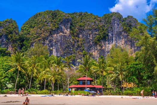 Bungalow house with red roof among coconut palms near the cliffs with people sunbathing and swimming in emerald water on Railay beach west, Ao Nang, Krabi, Thailand.