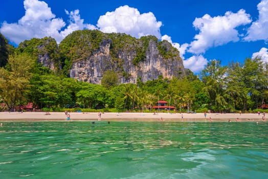 Bungalow house with red roof among coconut palms near the cliffs with people sunbathing and swimming in emerald water on Railay beach west, Ao Nang, Krabi, Thailand.