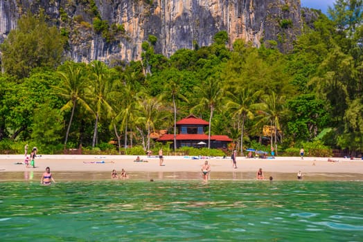 Bungalow house with red roof among coconut palms near the cliffs with people sunbathing and swimming in emerald water on Railay beach west, Ao Nang, Krabi, Thailand.