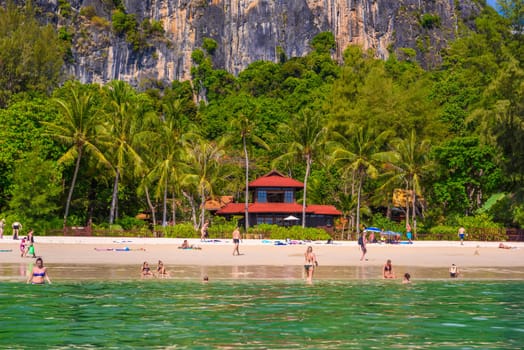 Bungalow house with red roof among coconut palms near the cliffs with people sunbathing and swimming in emerald water on Railay beach west, Ao Nang, Krabi, Thailand.