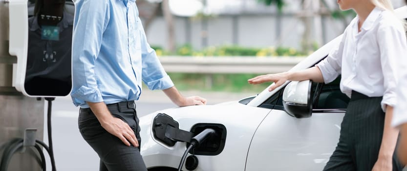 Progressive businessman and businesswoman with electric car parking and connected to public charging station before driving around city center. Eco friendly rechargeable car powered by clean energy.