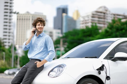 Progressive businessman talking on the phone, leaning on electric car recharging with public EV charging station, apartment condo residential building on the background as green city lifestyle.