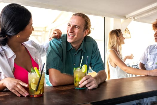 Happy young man enjoying mojito cocktail with girlfriend at a beach bar drinking. Young multiracial couple on a date. Lifestyle and summertime concept.
