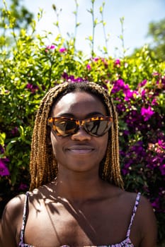 Vertical headshot portrait of African American young woman wearing cool sunglasses looking at camera. Flower and greenery background. Lifestyle.