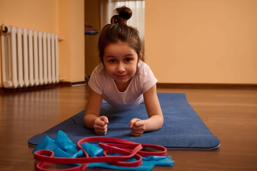 Front shot of a 5-6 years old Caucasian determined sporty little child girl, looking at camera, doing plank exercise on a blue fitness mat while working out, stretching body, practicing yoga at home