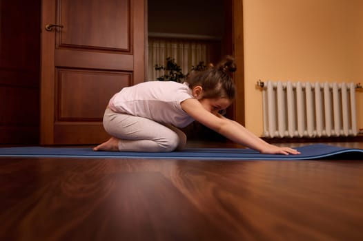 Side portrait of a Caucasian child, lovely baby girl in sportswear, stretching her body in child pose, practicing yoga on a blue fitness mat in a cozy wooden home interior. Healthy habits. Childhood