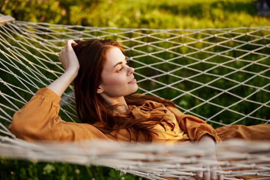 a happy woman is resting in a mesh hammock with her head resting on her hand, smiling happily looking away, enjoying a warm day in the rays of the setting sun, lying in an orange dress. High quality photo