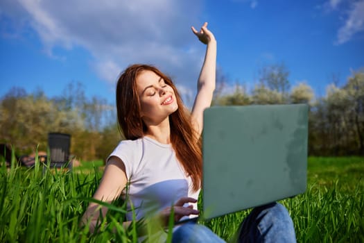 joyful woman works sitting in high grass behind a laptop raising her hand up. High quality photo