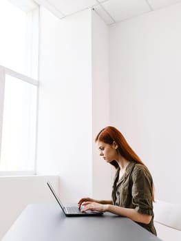 cute red-haired woman sitting at a table and working at a laptop looking seriously at the monitor. High quality photo