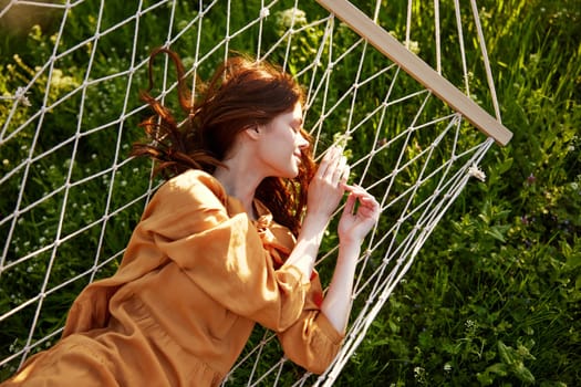 a beautiful, elegant woman lies in a long orange dress on a mesh hammock resting in nature, illuminated by the warm sunset light. High quality photo