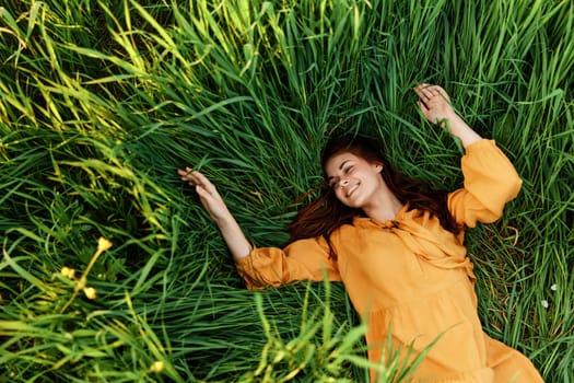 a relaxed red-haired woman enjoys summer lying in the tall green grass in a long orange dress smiling happily with her eyes closed. High quality photo