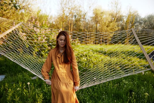 a beautiful, elegant woman is standing in a long orange dress leaning on a hammock smiling pleasantly. High quality photo