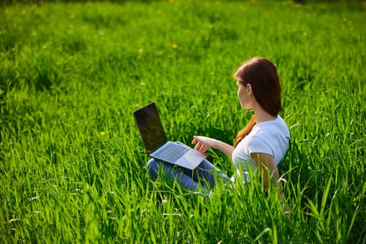 a woman in a light T-shirt works at a laptop while sitting in high green grass. High quality photo