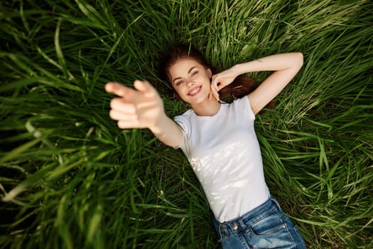 A happy girl in a white t-shirt stretches her arms up and lies on the green grass. High quality photo