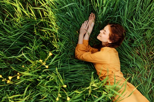 a relaxed red-haired woman enjoys summer lying in the tall green grass in a long orange dress smiling happily looking away. High quality photo