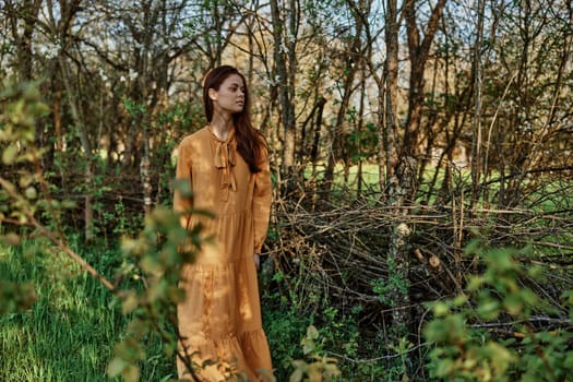 a beautiful, slender woman with long hair walks in the shade near the trees, dressed in a long orange dress, enjoying the weather and the weekend. The theme of privacy with nature. High quality photo