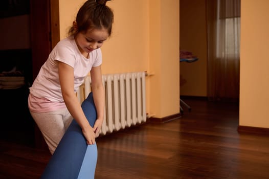 Caucasian adorable smiling little child girl in sportswear, unfolding blue fitness mat before working out at home interior. Healthy habits. Active lifestyle concepts. Yoga practice. Leisure activity