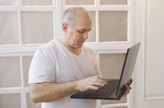 A man holds a laptop in his hands, stands at home and communicates in social networks, writes a message.