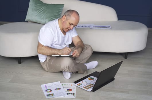 A man sits on the floor, works on a laptop, studies reporting documents, charts and graphs, makes notes in a notebook.