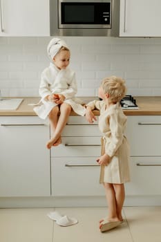 A girl is sitting in the kitchen and a boy is standing in dressing gowns, they are talking.