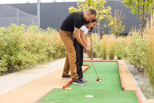 Sharing with golf experience. Cheerful young man teaching his daughter to play mini golf at the day time. Concept of friendly family.