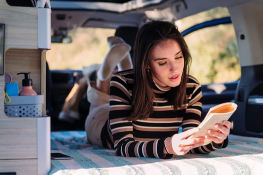 young caucasian woman relaxing in camper van reading a book, concept of road trip adventure and van life relaxation