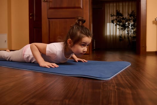 Determined little girl doing push ups, four limbed staff exercise, staying in plank on a blue fitness mat while practicing stretching and yoga indoors. Healthy habits and active lifestyle concept