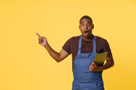 Amazed young African American cook in blue apron, searching for recipes online on tablet PC. Cook holding tablet in one hand and gesturing towards the copy space on bright yellow background with other hand. . High quality photo