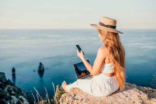 Successful business woman in yellow hat working on laptop by the sea. Pretty lady typing on computer at summer day outdoors. Freelance, travel and holidays concept.