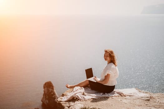 Successful business woman in yellow hat working on laptop by the sea. Pretty lady typing on computer at summer day outdoors. Freelance, travel and holidays concept.