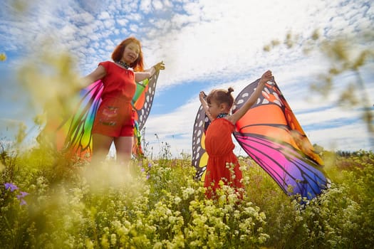 Happy female family with red haired mother and daughter with bright butterfly wings having fun on green and yellow meadow full of grass and flowers in sunny summer day. Concept family love