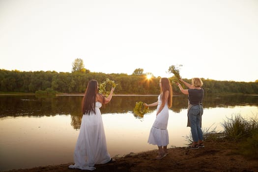 Three female girls in white sundresses and with flower wreaths on the riverbank at sunset. The feast of Ivan Kupala and national clothes and traditions of throwing wreath into the water