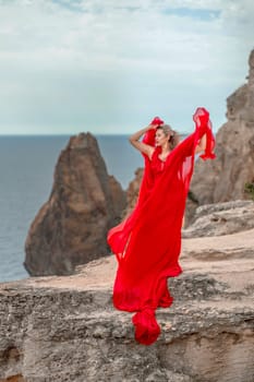 A woman in a red silk dress stands by the ocean, with mountains in the background, as her dress sways in the breeze