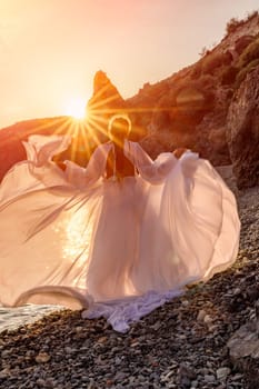 Mysterious woman silhouette long hair walks on the beach ocean water, sea nymph wind listens to the wave. Throws up a long white dress, a divine sunset. Artistic photo from the back without a face.