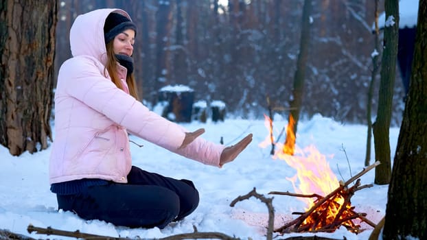 a large open-air fire used as part of a celebration, for burning trash, or as a signal. Young woman on a winter picnic in the forest warming up near the fire.