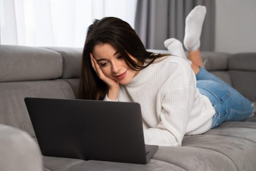 Young woman enjoys chat with friends via modern Notebook. Satisfied lady lies on couch in living room using modern laptop for online communication