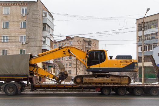 A heavy modern excavator stands on a low trailer platform along the rear ramp. Instead of a bucket, its boom has a tooth for loosening hard and frozen soil.Technique on a city street.