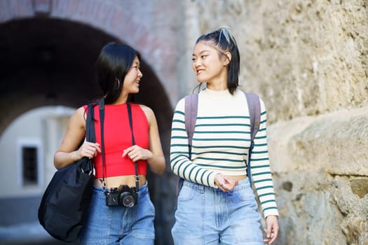 Happy young Asian female tourists in casual clothes with bags and photo camera looking at each other while strolling together near rough wall