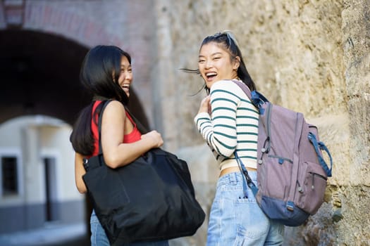 Cheerful Asian female tourists with belongings looking over shoulder at camera with backpack while standing near stone wall of building