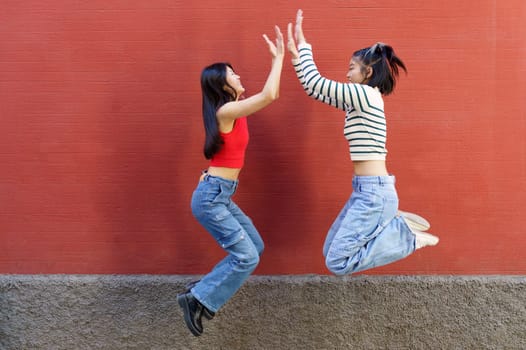 Full body side view of cheerful young Asian female friends jumping up and giving high five to each other against red wall