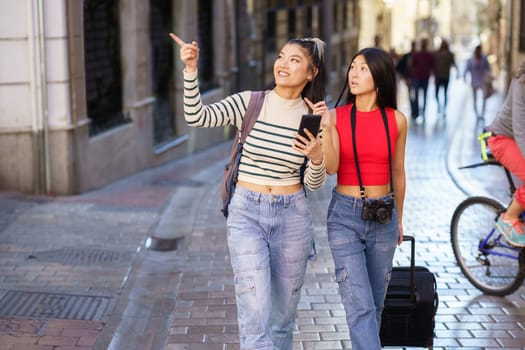 Young positive Asian girlfriends with backpack and suitcase walking on street of Granada and pointing at sightseeing while using cellphone during journey