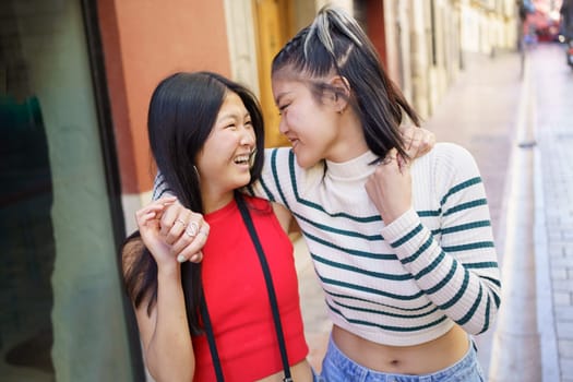 From above of stylish laughing ethnic female tourists walking on paved street in old town and hugging while looking at each other