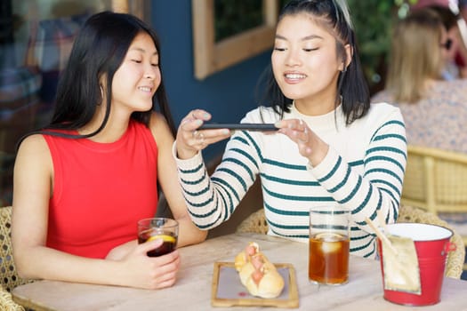 Smiling young Asian female friends, in casual clothes sitting at table with cold drinks and taking picture of tasty food on smartphone while spending time together in cafe