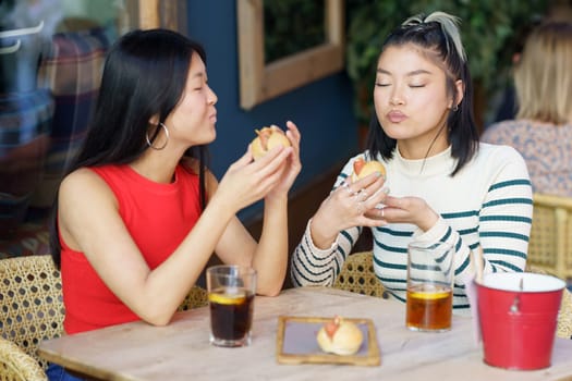 Delighted young Asian female friends in casual wear eating delicious food with closed eyes while sitting at table with cold beverages in cafe together